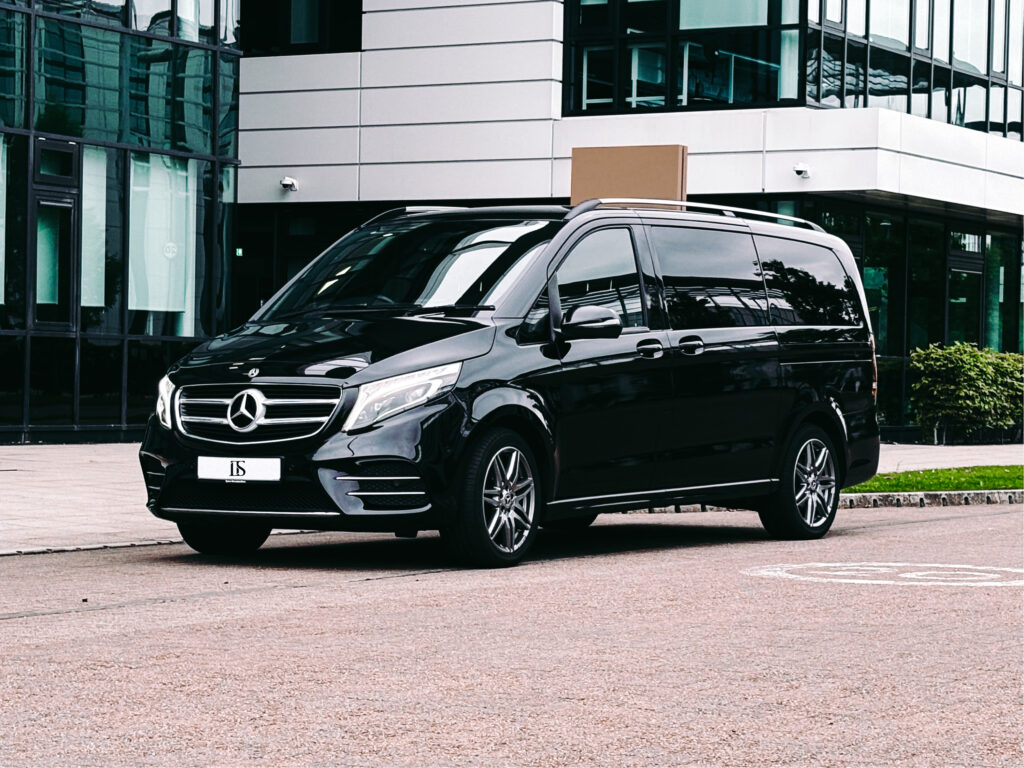 Image features Mercedes V-Class chauffeur driven vehicle. It is a black van against the backdrop of corporate buildings.